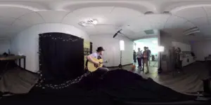 A musician playing guitar in a studio setting with crew members and lighting equipment in the background.