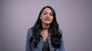 Woman with long black hair wearing a denim jacket and polka-dot top, smiling against a light gray background.