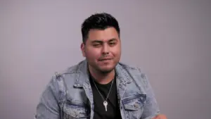 Man in a denim jacket and black shirt smiling at the camera against a neutral background, exuding confidence and warmth.