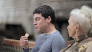 Young man enjoying a cookie while sitting beside a gray-haired woman in a relaxed outdoor setting.