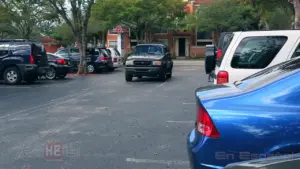 A pickup truck navigating a parking lot filled with various vehicles and lush trees in the background.