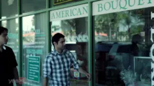 Man in plaid shirt gazes at flower shop display in a storefront, highlighting vibrant floral arrangements and signage.