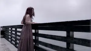 A woman in a long dress gazes pensively over a wooden pier, with calm water and a cloudy sky in the background.