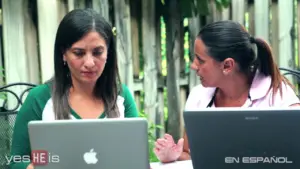 Two women working together on laptops outdoors, engaged in discussion, with a wooden fence in the background.