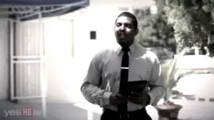 Man in a suit holding a tablet, standing outdoors near a building with trees and a blue awning in the background.