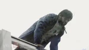 Man in a blue jacket leaning over an edge, set against a cloudy sky, conveying a sense of tension and urgency.