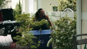 Man sitting on a blue cooler, laughing joyfully, surrounded by greenery and a pickup truck in a sunny outdoor setting.