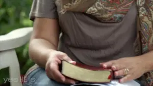 Person holding a closed book while sitting outdoors, wearing a patterned scarf and casual clothing.