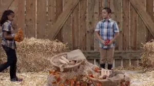 Children in casual clothes at a rustic setting with straw, baskets of vegetables, and autumn decorations.