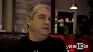 Man with gray hair sitting in a restaurant, looking surprised, with a blurred floral arrangement in the background.