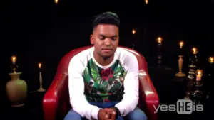 A young man sitting thoughtfully in a red chair, with soft lights and dark background enhancing the intimate atmosphere.