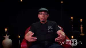 Man in a black t-shirt sitting on a red chair, discussing personal experiences in a dimly lit studio setting.