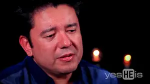 Close-up of a man with dark hair, expressing emotion, illuminated by soft candlelight in a contemplative setting.