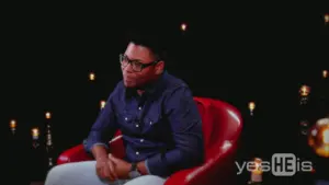 Man in glasses sitting in a red chair against a dark background with soft lights, engaged in conversation.