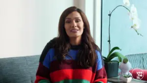 Woman in colorful striped sweater smiling, sitting beside a large window with a plant and modern decor in background.