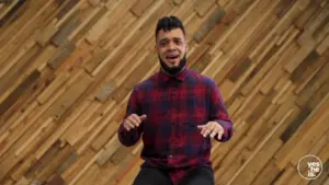 Man in a checkered shirt sitting against a textured wooden backdrop, smiling and engaging with the audience.