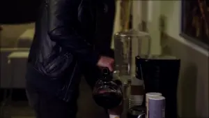 Person in a leather jacket pouring coffee from a carafe into a cup near a coffee maker and containers in a dimly lit kitchen.