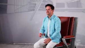 Man in a blue checkered shirt sitting on a stylish chair against a minimalist background.