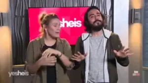 Two presenters engage in a lively conversation in front of a colorful backdrop featuring the logo "yes she's."