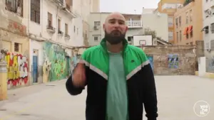 Man wearing a green jacket stands in a graffiti-filled basketball court, gesturing confidently in an urban setting.