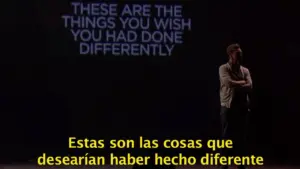 A performer stands on stage with projected text reflecting on regrets and life choices in English and Spanish.