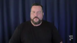 Man with a beard wearing a black shirt, sitting in front of a dark blue curtain, appearing thoughtful.