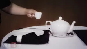 Hand pouring tea from a white teapot into a small cup, set on a neatly arranged table with black and white linens.