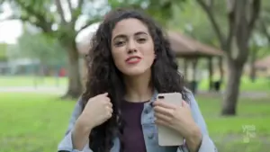 Young woman with curly hair holding a phone, smiling in a green park setting with trees and a gazebo in the background.