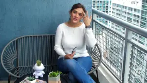 Woman sitting on a balcony bench, holding a phone, with small plants and a cityscape in the background.