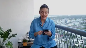 Woman sitting on a balcony, smiling while using a smartphone, with a cityscape view in the background.