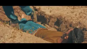 Man lying in a shallow grave as another person digs around him in a desolate landscape, showcasing tension and drama.