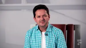 Man in a green plaid shirt sitting in a modern chair, smiling against a minimalistic background.