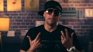 Man in sunglasses and cap gesturing, with a brick wall and shelves in the background, showcasing a confident vibe.