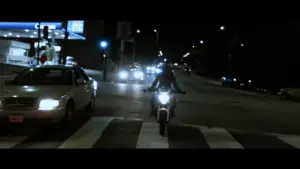 Motorcyclist riding at night on a street crossing, with cars and city lights in the background.