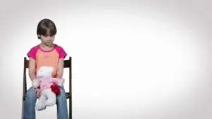 Young girl in a colorful shirt sitting on a chair, holding a stuffed animal, with a simple background.