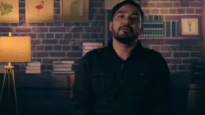 Man in a black shirt sitting against a brick wall with books and artwork, looking thoughtfully at the camera.