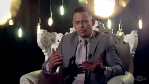 Man in a suit sitting on a decorative chair, gesturing thoughtfully, with soft lights in the background.