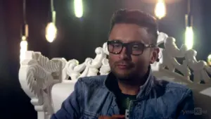 Man in glasses wearing a denim jacket, seated on an ornate chair with warm, glowing pendant lights in the background.