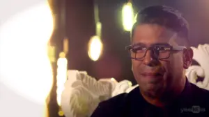 Emotional man with glasses, sitting on a decorative chair, illuminated by soft lights in an intimate setting.