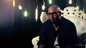 Bald man in glasses sitting on a luxurious white chair with colorful pendant lights in a dimly lit background.