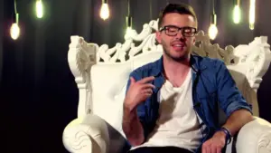 Young man in glasses sitting on an ornate white chair, gesturing while speaking, with hanging lights in the background.