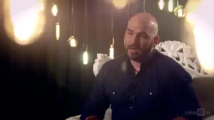 Man with a beard sitting in a well-lit room, speaking against a backdrop of hanging light fixtures.