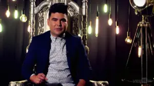 Man seated on an ornate throne with stylish light fixtures in the background, wearing a patterned shirt and blazer.