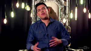 A man sitting confidently on an ornate throne, surrounded by hanging light bulbs, speaking engagingly to the camera.