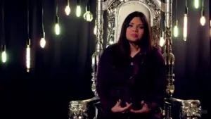 Woman seated on an ornate throne with hanging light bulbs in a dimly lit background, emphasizing a regal atmosphere.