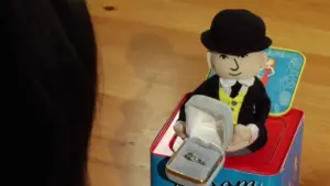 A plush toy character in a tuxedo presents an engagement ring in a small box during a playful moment.