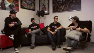 Four friends in black t-shirts engage in conversation on a couch, surrounded by posters and decor in a casual setting.