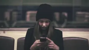 Young woman in a beanie sitting alone in a subway, focused on her smartphone, surrounded by dimly lit train interior.