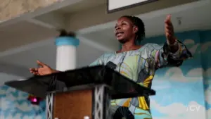 Woman speaking passionately at a podium in a colorful dress, engaged in a lively presentation at a conference or event.