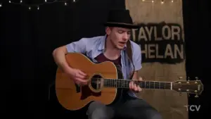 Musician in a black hat passionately playing an acoustic guitar on stage, with light decorations in the background.